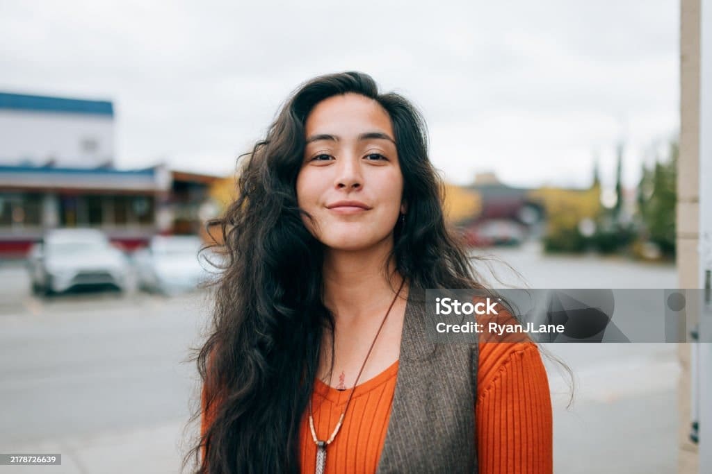 Une jeune femme aux longs cheveux bruns ondulés sourit doucement à l'extérieur. Vêtue d'un haut orange et d'un gilet marron, elle se tient devant une rue et des immeubles flous, par une journée nuageuse.