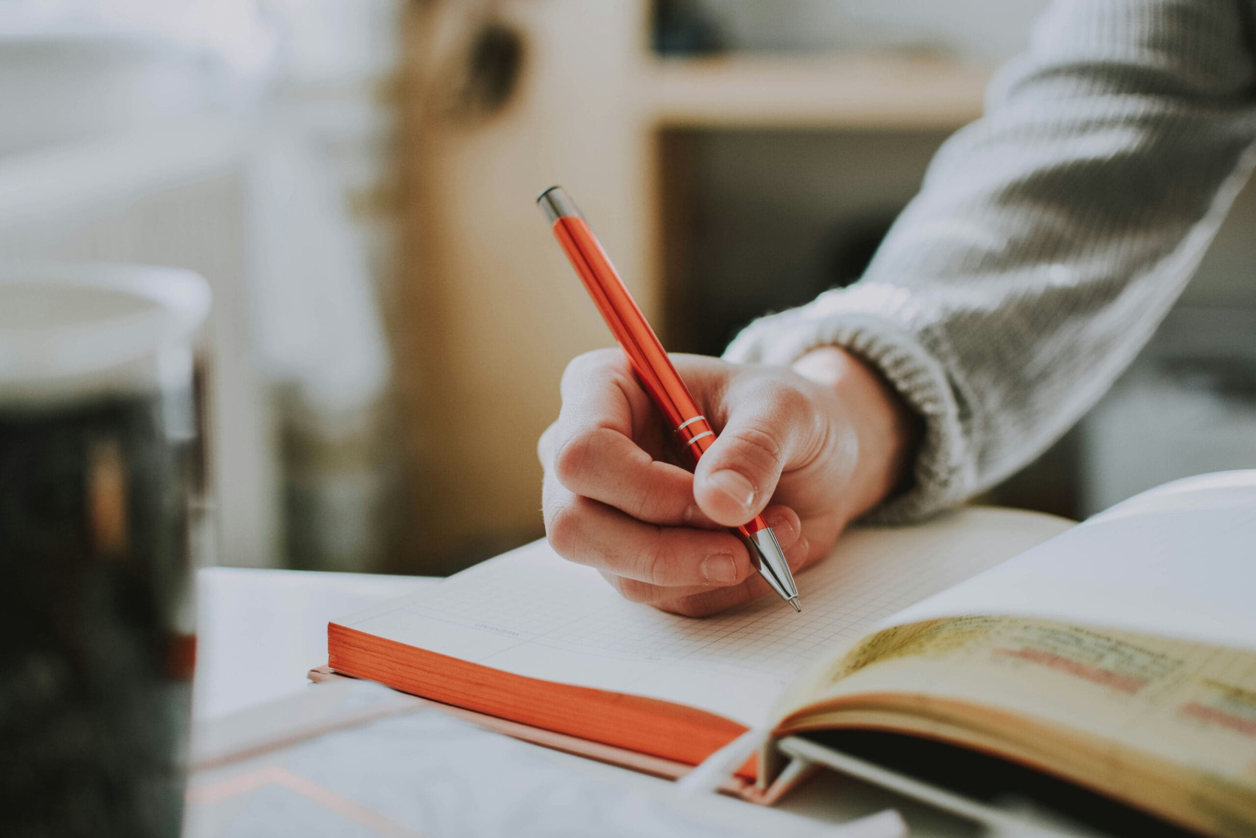 Une personne portant une manche grise écrit dans un cahier avec un stylo rouge. Le cahier est ouvert sur un bureau ; on aperçoit une tasse et un autre livre ouvert dans l'arrière-plan flou.