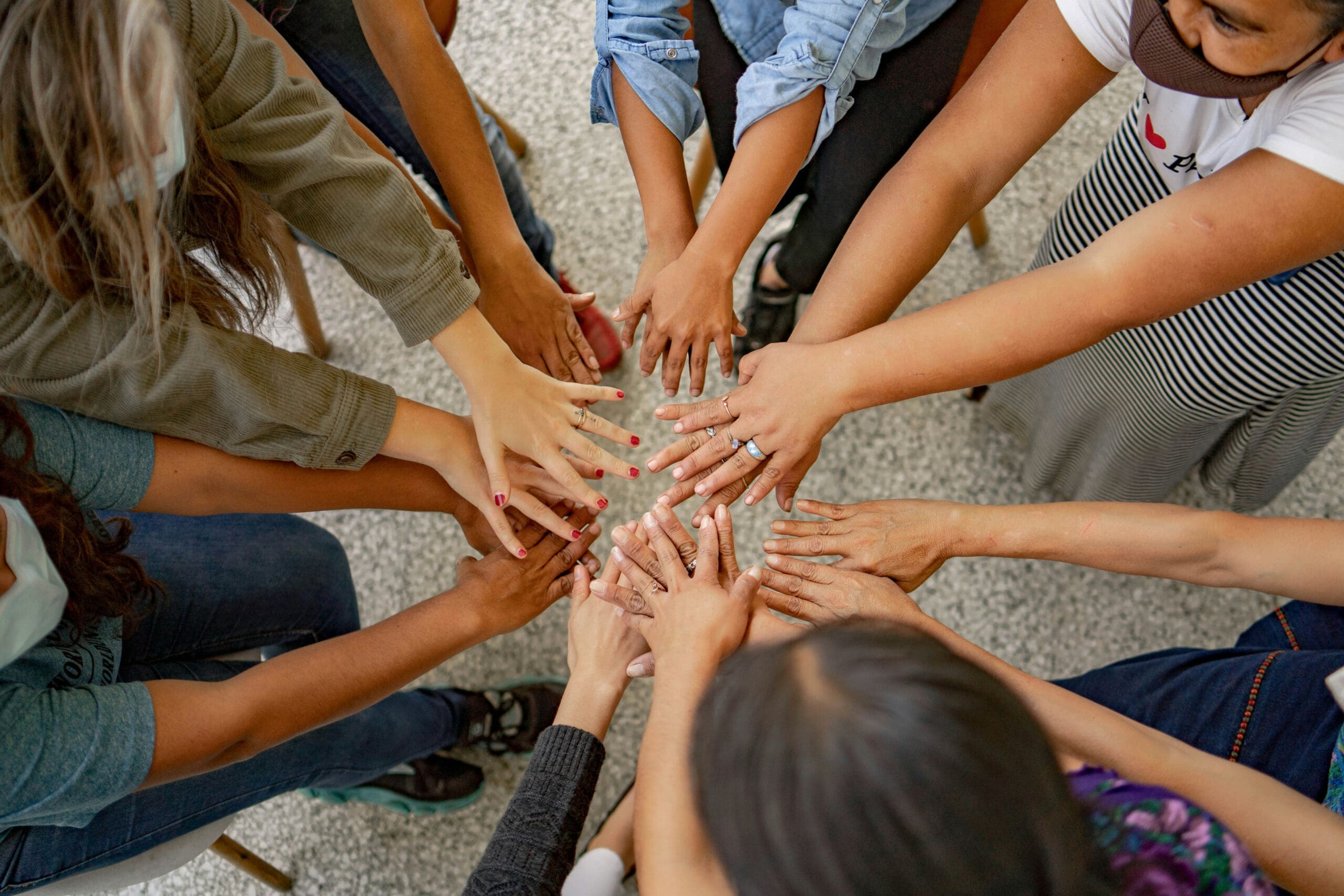 Un groupe de personnes assises en cercle tendent les mains vers le centre, en se touchant du bout des doigts, symbolisant ainsi l'unité et la solidarité.